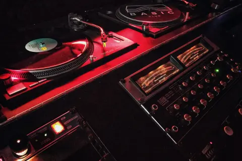 Giradischi e mixer in un locale notturno, illuminati da luci rosse. Il logo "BASIC CLUB" è visibile su uno dei giradischi.
