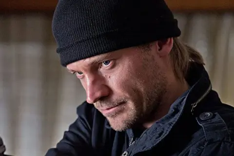 En man med svart mössa och skäggstubb, kanske en karaktär från filmer och tv-program med Jo Nesbø.