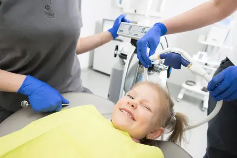 Uśmiechnięta dziewczynka na fotelu dentystycznym, stomatolog przygotowuje sprzęt.