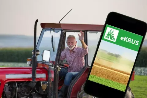 Uśmiechnięty rolnik w traktorze macha do kamery, obok smartfon z aplikacją eKRUS, która ułatwia zarządzanie prawem do emerytury rolniczej według nowych zasad.