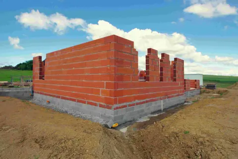 Budowa domu 100m2 w toku. Czerwone ściany z pustaków, fundamenty z bloczków betonowych, niebo z chmurami.