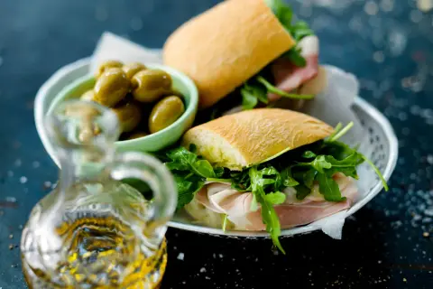 Två halvor av en ciabatta macka med prosciutto och ruccola, serverade med gröna oliver och en karaff olivolja.