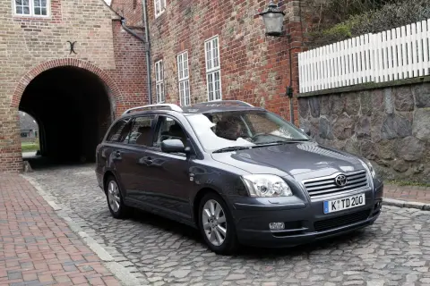 Szary Toyota Avensis T25 2.0 D4D na brukowanej uliczce, obok ceglanego budynku z łukiem.