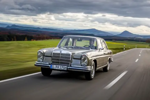 En klassisk Mercedes 60-tal glider fram på en slingrande landsväg med böljande gröna fält och dramatiska berg i bakgrunden.