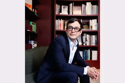 A man in a suit and glasses sits in front of bookshelves, perhaps at the David Lewis Gallery, looking thoughtfully to the side.