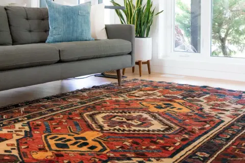 Ein detailreicher perserteppich mit roten und blauen Mustern liegt vor einem grauen Sofa. Tipps zum perserteppich reinigen.