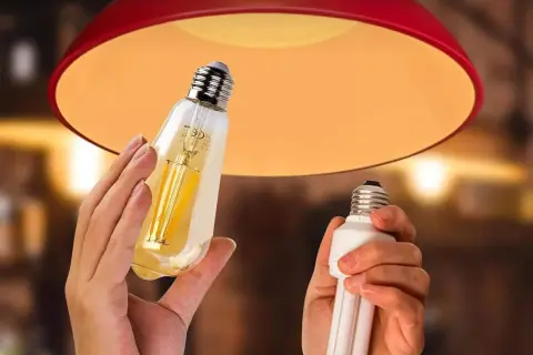 Une ampoule connectée orange, au design vintage, est tenue à côté d'une ampoule fluocompacte, sous un abat-jour rouge.