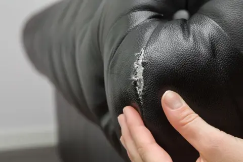 Main d'une personne examinant un accroc sur un fauteuil en cuir noir, montrant la nécessité de réparer le cuir craquelé.