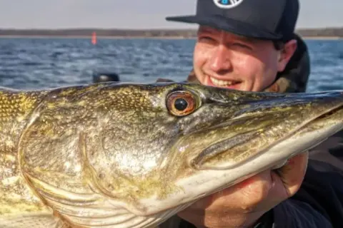 Glücklicher Angler hält stolz einen großen Hecht, gefangen nach einem Gewitter. Das Wasser ist ruhig, die Sonne scheint.