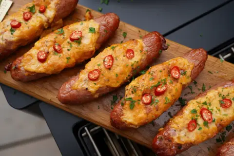 Gerichte mit grober Bratwurst, überbacken mit Käse und garniert mit Chili und Petersilie, liegen auf einem Holzbrett.