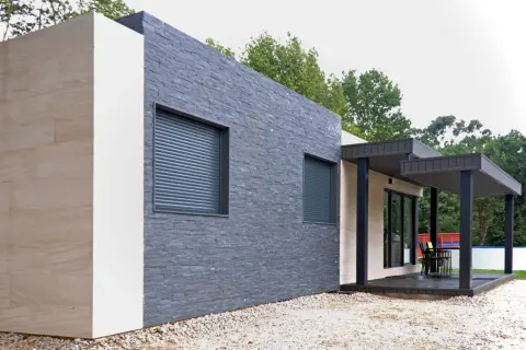 Muros exteriores de casas modernas con texturas de piedra y acabados claros. Ventanas con persianas enrollables y un porche con mobiliario.