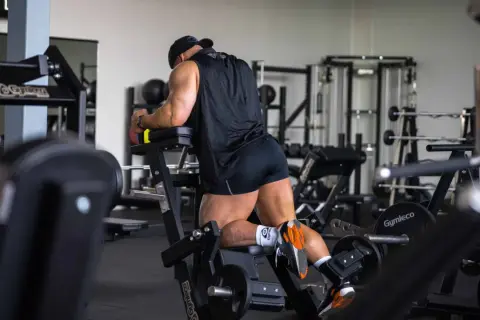 Man utför stående lårcurl på ett gym. Han har en svart tröja och shorts, och tränar sina baksida lår.