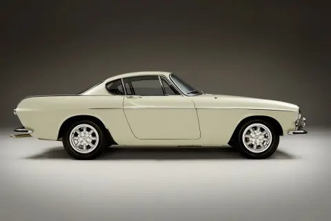 Ein cremefarbener Volvo P1800 Coupé steht im Profil, seine eleganten Linien und Chromdetails glänzen.