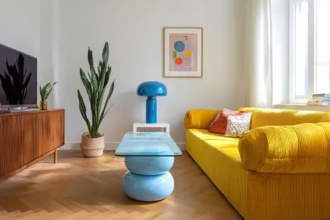 Jasny salon retro z żółtą kanapą, niebieską lampą i stolikiem kawowym. Drewniana komoda i duża roślina dopełniają przytulną atmosferę.