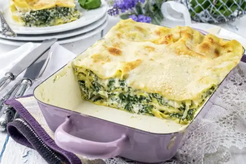 Pyszne lasagne ze szpinakiem w fioletowym naczyniu, gotowe do podania. Obok sztućce i dekoracyjne kwiaty.