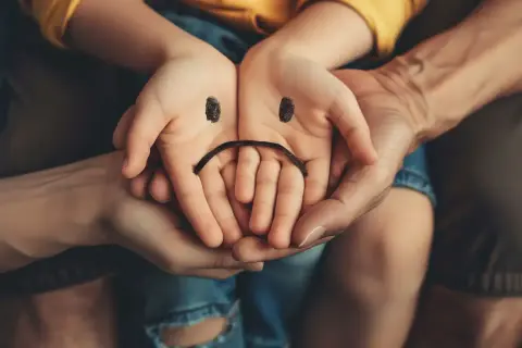 Dziecięce dłonie z namalowaną smutną buźką, otoczone dłońmi dorosłych, symbolizują zaburzone relacje w rodzinie i poczucie smutku.