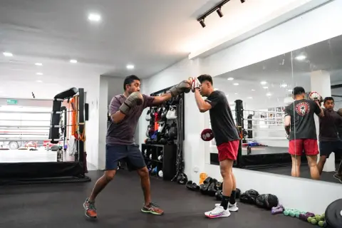 Two men practice boxing conditioning exercises in a gym. One man throws a punch while the other holds pads.