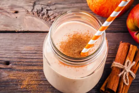 Pyszny drink szarlotka w słoiku, posypany cynamonem, obok jabłka i laski cynamonu na drewnianym tle.