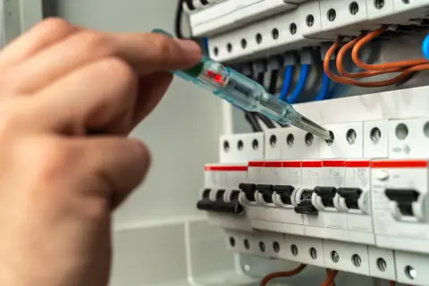 Dłoń z próbnikiem napięcia sprawdza zabezpieczenie skrzynki elektrycznej. Czerwona lampka próbnika świeci.