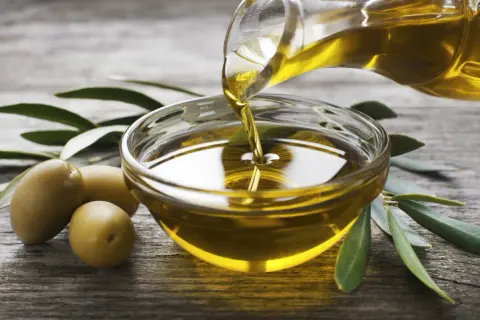 Olio evo l'extravergine, dorato e puro, viene versato in una ciotola di vetro. Olive e foglie di ulivo completano la scena.