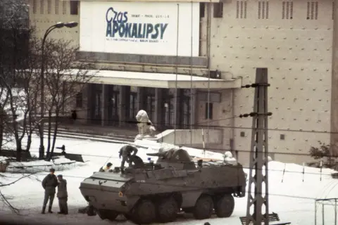 Wóz bojowy na śniegu, ludzie w mundurach, budynek z napisem "Czas Apokalipsy". Atmosfera przypomina stan wojenny.