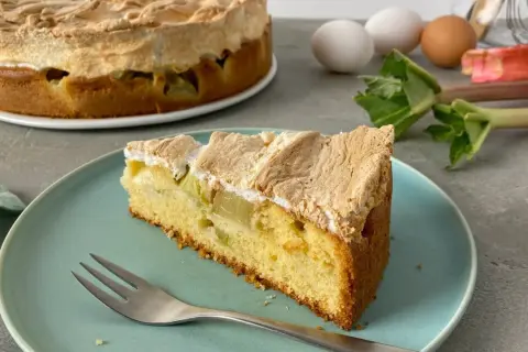 Ein saftiges Stück Rhabarber Baiser Kuchen mit einer luftigen Baiserhaube liegt auf einem blauen Teller. Im Hintergrund ein ganzer Kuchen und frischer Rhabarber.