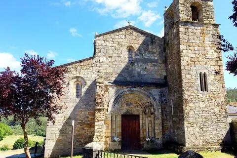 La catedral de Foz, con su torre románica y fachada de piedra, se alza bajo un cielo azul con nubes. Un árbol de hojas rojizas añade color.