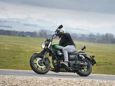 Motocyklista na zielonym chopperze UM Renegade 125 przemierza malowniczą drogę wśród zielonych pól i gór.