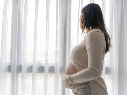 Kobieta w ciąży stoi przy oknie, patrząc na zewnątrz. Jej dłonie delikatnie obejmują brzuch, a w jej postawie można wyczuć smutek, sugerujący depresję ciążową.