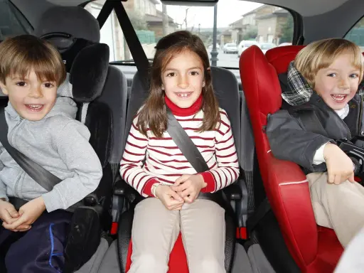 Tres niños sonríen en sus asientos de coche. El niño de la izquierda, con sudadera gris, parece preguntarse cuándo puede ir un niño delante.