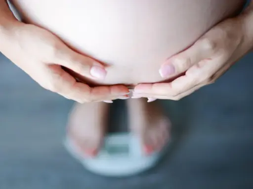 Dłonie obejmujące brzuch ciężarnej, stojącej na wadze. Schudłam w ciąży 10 kg, co widać na wadze.