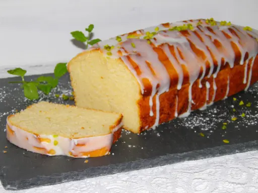 Ein saftiger mallorquinischer Zitronenkuchen mit Zuckerguss und Pistazien, daneben ein angeschnittenes Stück.