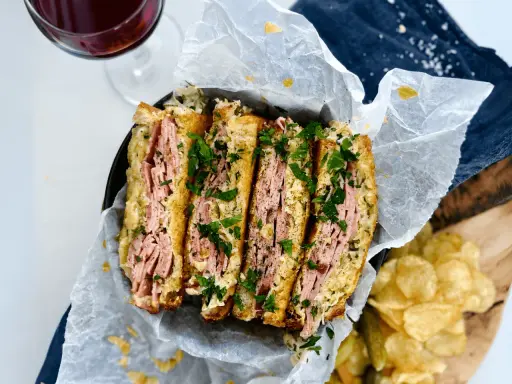 En saftig reuben sandwich, skuren i fyra bitar och toppad med färsk persilja. Serveras med krispiga potatischips och en glas rödvin.