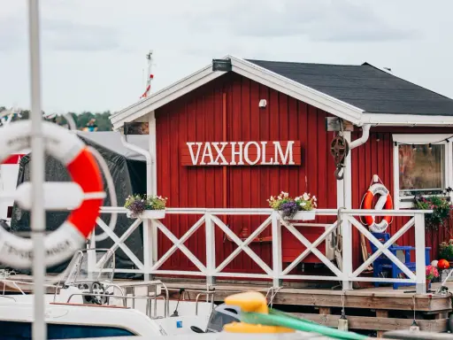 Rött hus med texten VAXHOLM vid vattnet. En livboj hänger på väggen. Vaxholms kastell skymtar i bakgrunden.