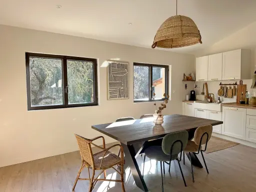 Comedor y cocina luminosos con mesa de madera, sillas de mimbre y tapizadas, y ventanas de cristal térmico que dejan ver el exterior.