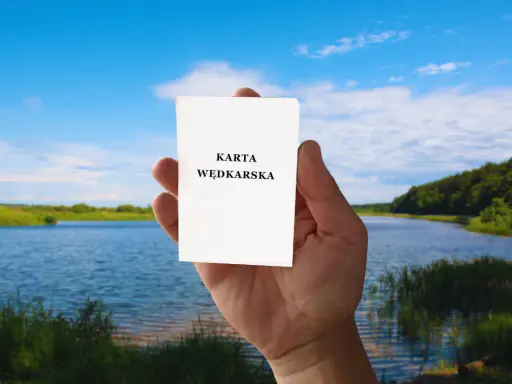 Dłoń trzyma białą kartkę z napisem "KARTA WĘDKARSKA" na tle jeziora i lasu.