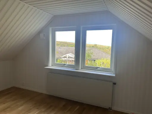 Vitt snedtak med panel och ett fönster med utsikt över träd. Gardiner saknas, men ett element finns under fönstret.