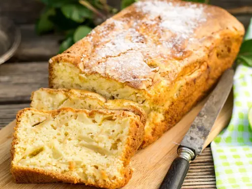 Szybkie ciasto z jabłkami, posypane cukrem pudrem, pokrojone na kromki, leży na desce obok jabłek i noża.