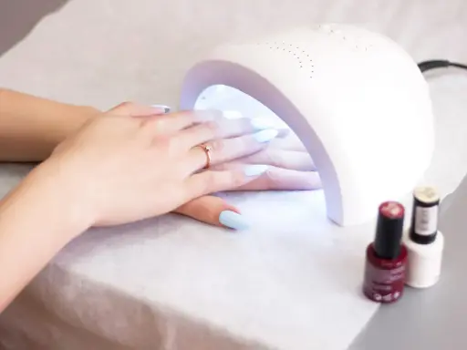 Dłonie z jasnoniebieskimi paznokciami pod lampą UV. W tle stoją buteleczki z lakierami. Lampa UV w ciąży - delikatna pielęgnacja.