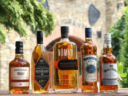 Pięć butelek szkockiej whisky: Edradour, Antiquary, Nomad, Claymore i Whyte & Mackay, stojących na drewnianym stole.