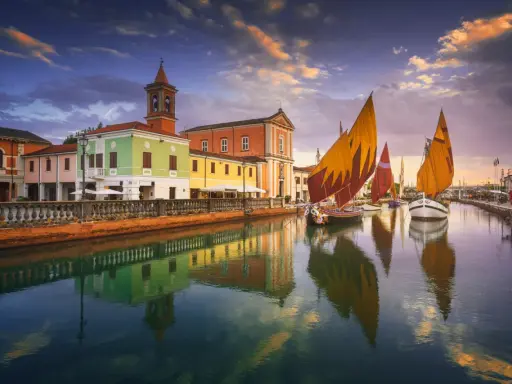 Navi a vela colorate navigano sul canale di Cesenatico al tramonto, con edifici vivaci e riflessi sull'acqua. Meglio Cervia o Cesenatico? Cesenatico offre questo spettacolo.