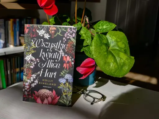 Okładka książki "Wszystkie kwiaty Alice Hart" z motywem roślinnym, obok cyklamen i okulary.