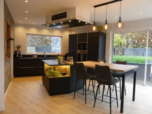 Cuisine noir et bois, mur gris texturé. Îlot central avec éclairage, table haute et tabourets. Fenêtre donnant sur jardin.