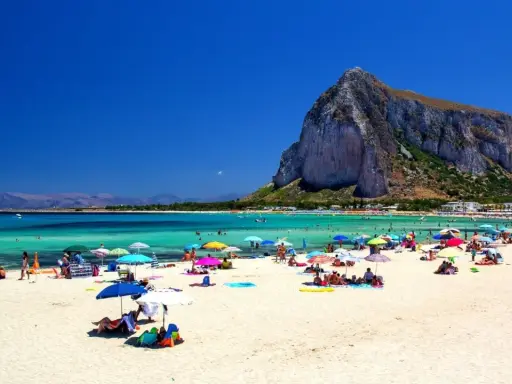 Spiaggia affollata con ombrelloni colorati, gente che si gode le vacanze al mare con bambini, acque turchesi e una montagna imponente sullo sfondo.