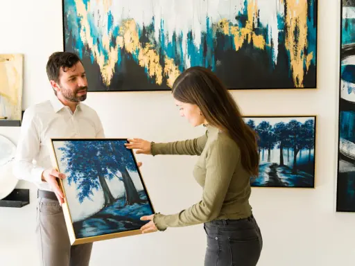 A man and woman admire a painting of blue trees at an art exhibition.