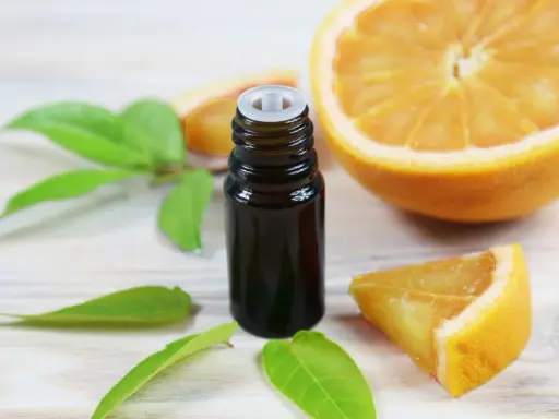 Flacon d'huile essentielle de citrus sinensis, entouré de feuilles vertes et de morceaux d'orange fraîche sur fond de bois clair.