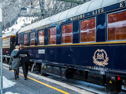 Ett par omfamnar varandra vid en snötäckt station, redo för en resa med Orientexpressen. Tågets mörkblå vagnar med gyllene detaljer vittnar om dess klassiska orientexpressen rutt.