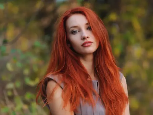 Eine Frau mit leuchtend roten Haaren steht im Freien. Ihre langen, feurigen Haare umrahmen ihr Gesicht, während sie in die Kamera blickt. Die Natur im Hintergrund ist unscharf und zeigt herbstliche Farben.