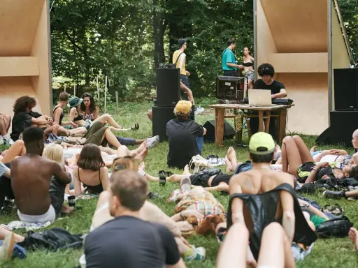 Un gruppo di persone rilassate su un prato in un **boutique festival**, con un DJ che suona musica in sottofondo.