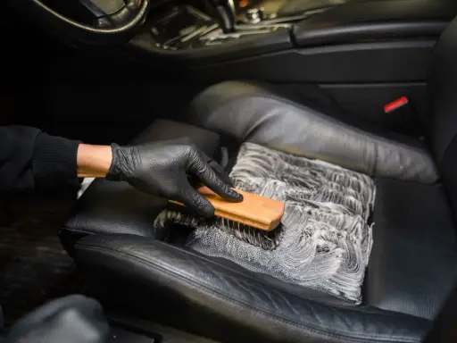 Mano enguantada cepilla asiento de cuero negro con espuma, mostrando cómo limpiar asientos de cuero para un coche impecable.